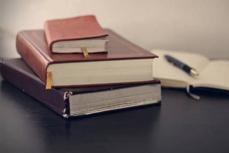 Três livros encadernados empilhados sobre uma mesa escura, ao lado de um caderno aberto com uma caneta tinteiro